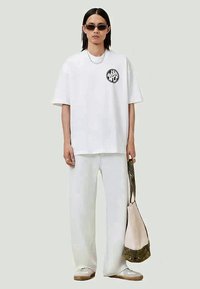 Pessoa vestindo uma t-shirt oversized branca e calças, óculos de sol escuros, segurando uma tote bag bege e verde, com ténis, em um fundo simples.