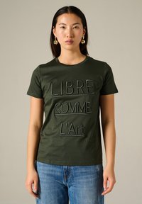 T-shirt en coton vert olive à manches courtes avec broderie du texte "LIBRE COMME L'Art" en noir. Encolure ronde, coupe décontractée.