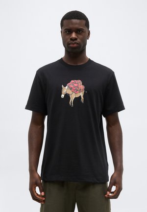 Uomo che indossa una maglietta nera con una stampa frontale di un asino che porta pacchi rossi, in piedi contro uno sfondo chiaro e uniforme.