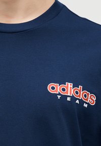 Μπλε μπλούζα ναυτικού τύπου με στρογγυλή λαιμόκοψη που φέρει κόκκινο και λευκό λογότυπο "adidas TEAM" στο στήθος.