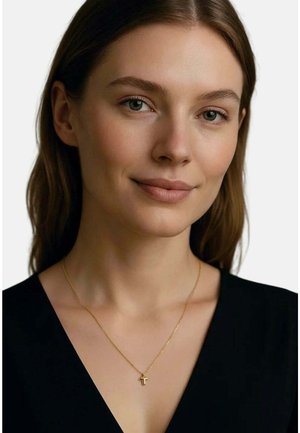 Junge Frau mit heller Haut und braunen Haaren, die ein schwarzes Oberteil und eine filigrane goldene Halskette mit kleinem Anhänger trägt.