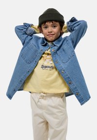 Veste en jean avec deux poches poitrine, de couleur bleue et boutons dorés, superposée à un t-shirt jaune clair avec un graphisme. Pantalons avec taille élastique blanche.