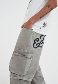 Light grey cargo pants with two large side pockets, featuring black graphic designs. Made from sturdy cotton material with a relaxed fit.