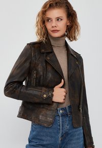 Giubbotto da motociclista in pelle marrone con finitura invecchiata, tasche con zip e revers con bottoni a pressione, indossato sopra un dolcevita beige.