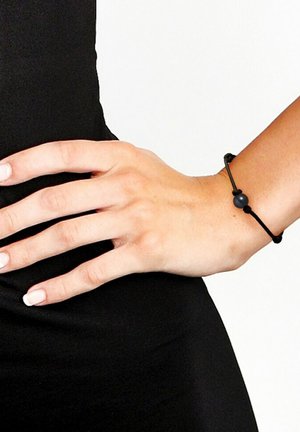 Bracelet en corde noir avec une perle sombre, design simple, longueur ajustable, porté sur une main. Texture lisse, style minimaliste.