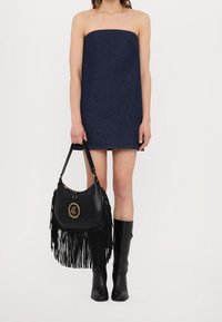 Borsa a tracolla in pelle nera con dettagli in frange e un accento del logo dorato, abbinata a un vestito strapless in denim scuro e stivali neri fino al ginocchio.