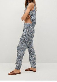 Vrouw in een mouwloze top met paisleypatroon en bijpassende losse broek, gecombineerd met bruine platte sandalen, zijwaarts staand met gekruiste armen.