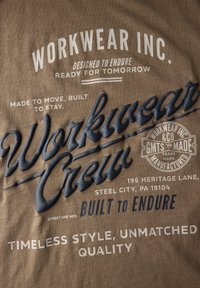 Hnědá látka s několika bílými a tmavě modrými potisky a ražbami textů o firmě Workwear Inc., včetně sloganů a adresy ve Steel City, PA.