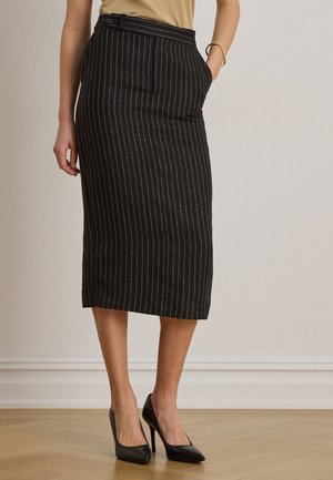 Femme portant une jupe crayon noire à fines rayures, un haut beige, des talons hauts noirs, debout avec une main dans la poche de la jupe contre un mur uni.
