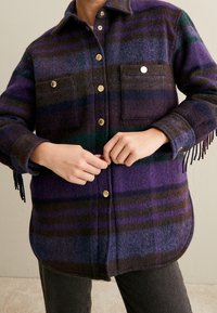 Camisa de lana a cuadros en morado, verde y marrón. Presenta dos bolsillos en el pecho, botones dorados y detalles de flecos en las mangas.