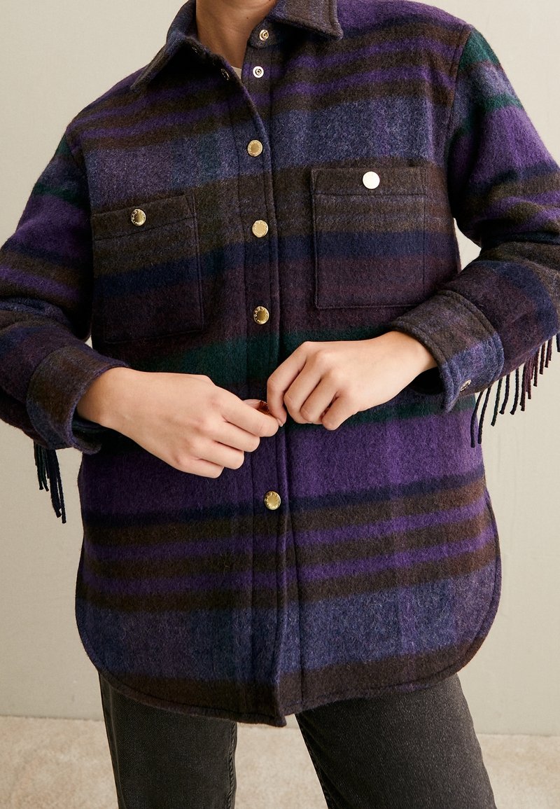 Camisa de lana a cuadros en morado, verde y marrón. Presenta dos bolsillos en el pecho, botones dorados y detalles de flecos en las mangas.