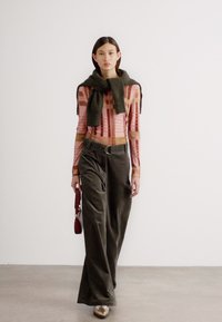 Modèle portant un haut rose à manches longues à motifs et un pantalon large vert olive, avec un pull vert foncé drapé autour des épaules.