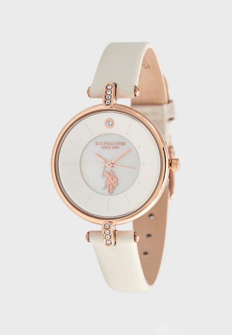 Reloj redondo con esfera blanca, caja de oro rosa y correa de cuero blanco. Presenta un logotipo, marcadores horarios y piedras decorativas cerca de la hebilla.