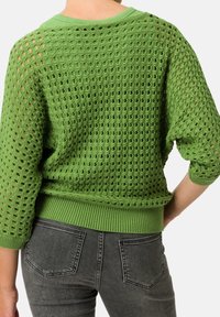 Persoon die een groene gehaakte trui met driekwartmouwen en verweerde zwarte spijkerbroek draagt, gezien van achteren.