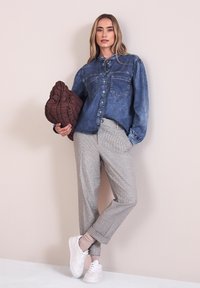 Femme portant une chemise en denim bleu, un pantalon gris à carreaux, des baskets blanches, tenant un sac matelassé marron, debout contre un mur pâle.