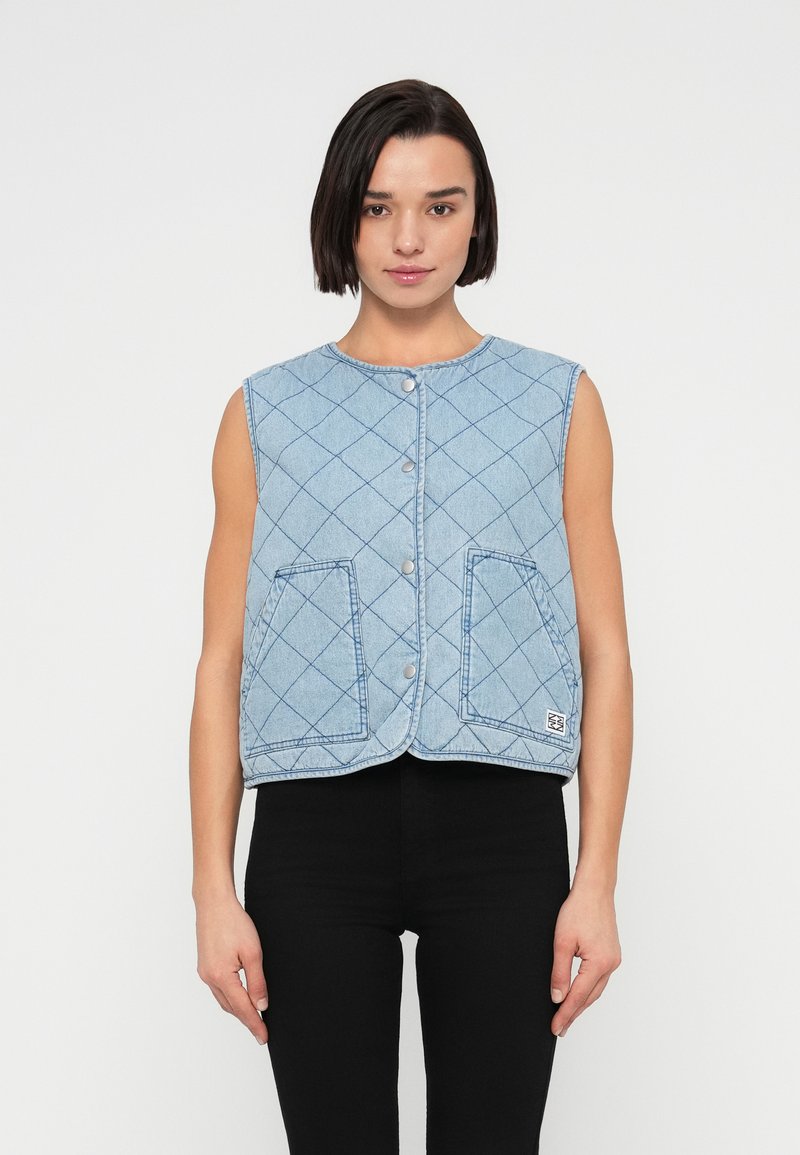 Καπιτονέ γιλέκο από τζιν σε ανοιχτό μπλε, με στρογγυλή λαιμόκοψη, μπροστινό κλείσιμο με κουμπιά και δύο μπροστινές τσέπες. Διαθέτει διακριτικό μοτίβο σε σχήμα ρόμβου.