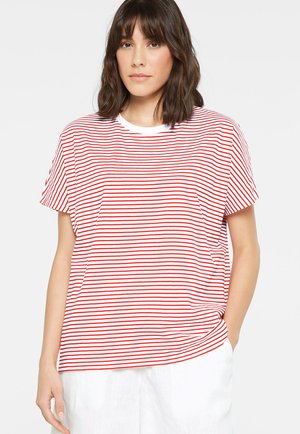 Frau mit schulterlangem dunklem Haar, die ein weißes T-Shirt mit dünnen roten horizontalen Streifen und weiße Hose trägt, Hand in der Tasche.