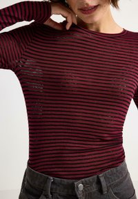 Frau in einem enganliegenden, langärmeligen, kastanienrot und schwarz gestreiften Shirt, einen Arm erhoben mit der Hand am Kopf, kombiniert mit dunklen, hochtaillierten Hosen.