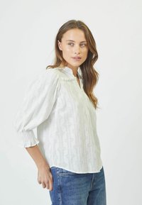 Camicia bianca con motivo testurizzato, colletto alla cinese e maniche a sbuffo. Abbinata a jeans di denim blu per un contrasto di colore e stile.