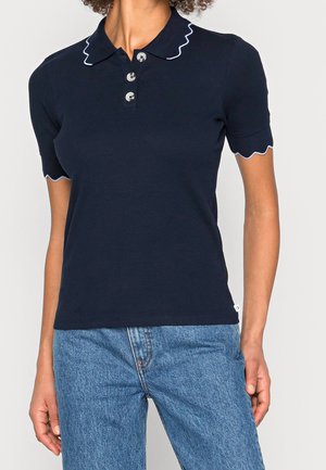 Mujer con polo de punto azul marino de manga corta y bordes blancos con ondas, y vaqueros azules, de pie con los brazos relajados.