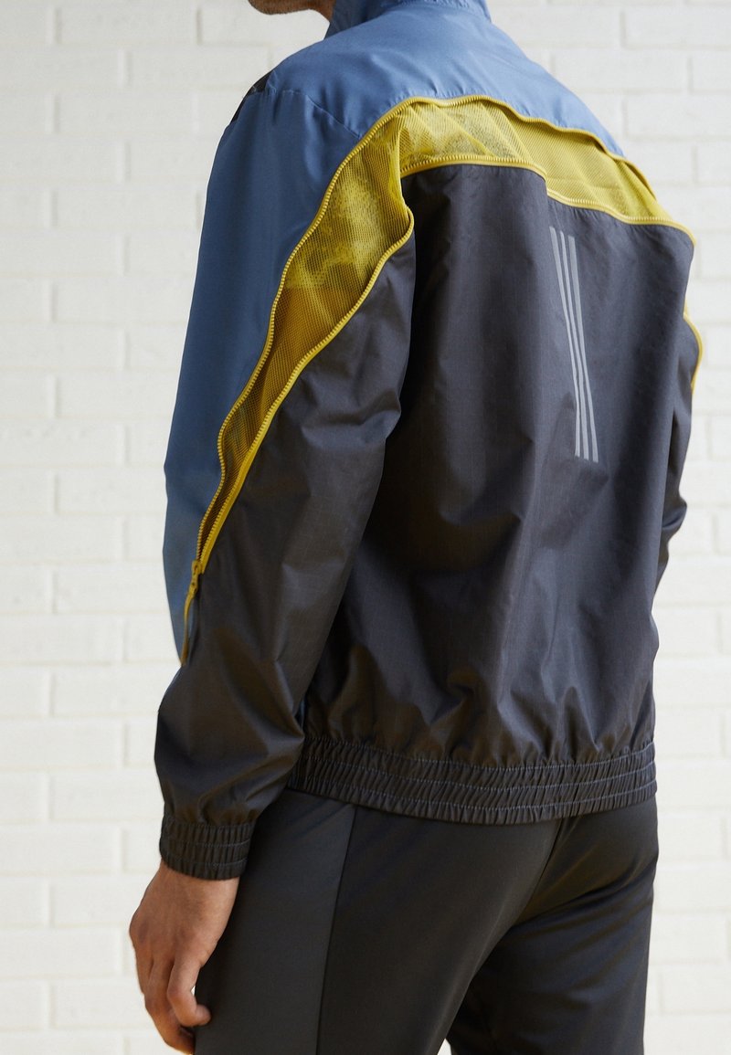 Windbreaker-Jacke in Blau, Schwarz und gelbem Mesh. Verfügt über eine Reißverschlusstasche, elastische Bündchen und reflektierende Streifen auf dem Rücken.