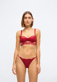 Sarkana me�žģīņu bralette ar polsterētiem kausiem, regulējamiem siksniņām un zelta detaļu akcentiem. Atbilstošs sarkans mežģīņu tanga apakšveļa ar delikātu dizainu.