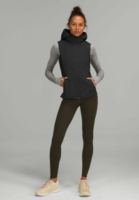 Gilet nero con cappuccio e zip frontale, abbinato a una maglietta grigia a maniche lunghe e leggings verde oliva. Scarpe da ginnastica chiare completano il look.