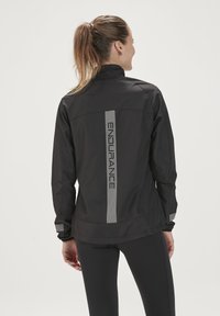 Zwarte lichtgewicht hardloopjas met een hoge kraag, reflecterend logo en strikingsdetails; beschikt over elastische manchetten en een iets gebogen zoom.