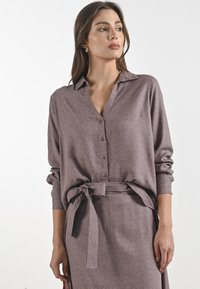 Set composto da camicia a maniche lunghe con colletto e gonna in malva con un sottile motivo geometrico. Presenta una cintura con fiocco e dettagli con bottoni. Texture liscia.