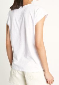 Bílá bavlněná tričko s načechranými krátkými rukávy a uvolněným střihem, kombinované s světlými béžovými šortkami. Tkanina je hladká a mírně texturovaná.