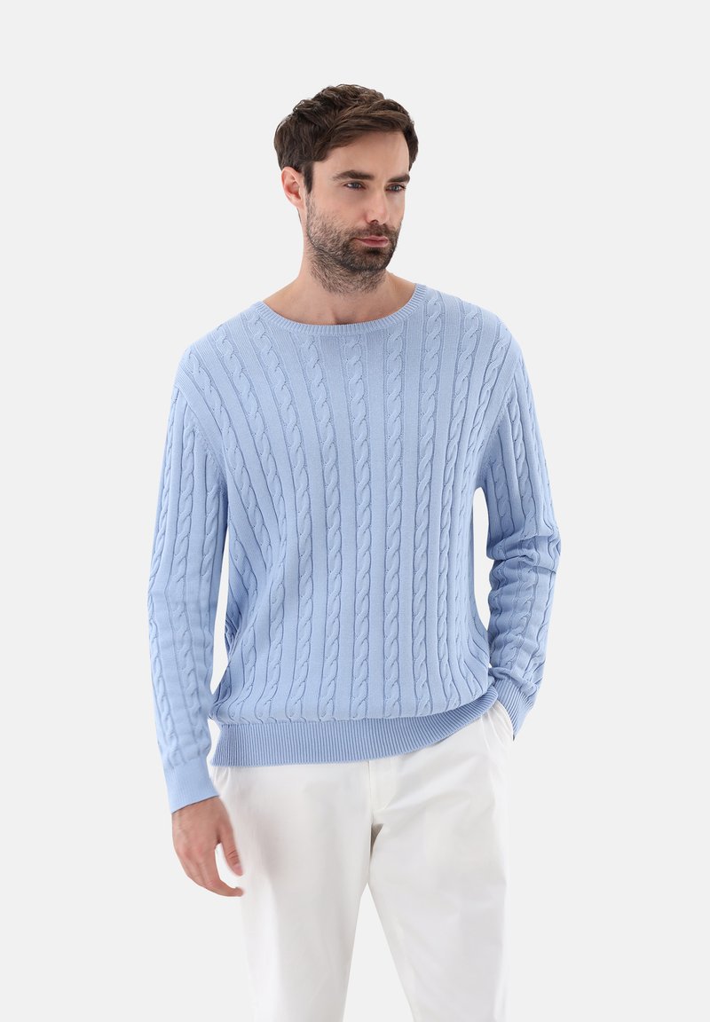 Hellblauer Kabelstrickpullover mit Rundhalsausschnitt und gerippten Bündchen, versehen mit einem strukturierten Muster und lockerer Passform. Kombiniert mit weißen Hosen.