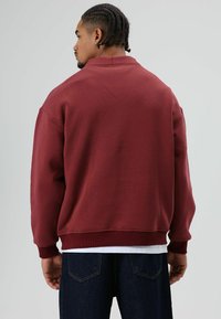 Bordeauxrode sweatshirt met een geribbelde kraag en manchetten, met een losse pasvorm. Draagt over een wit overhemd en gecombineerd met donkerblauwe spijkerbroek.