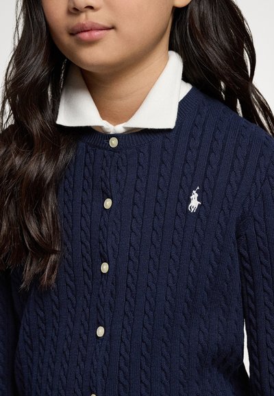 Cardigan azul-marinho de malha com padrão de trança, com fecho de botões brancos e um pequeno logotipo branco no peito esquerdo. Por baixo, é visível uma camisa branca com colarinho.