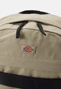 Barna vászon hátizsák fekete részletekkel, fekete cipzárral, két fekete pánttal és egy téglalap alakú Dickies logóval.
