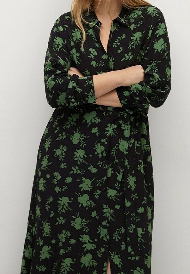 Femme les bras croisés portant une robe noire à boutons avec un motif floral vert et une ceinture à la taille.