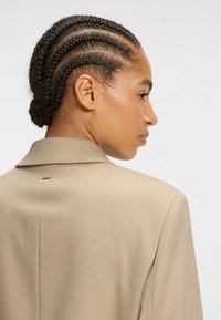 Beige uldblazer med en struktureret krave, synlige sømdetaljer og et syet mærke på bagsiden. Håret stylet i flere fletninger.