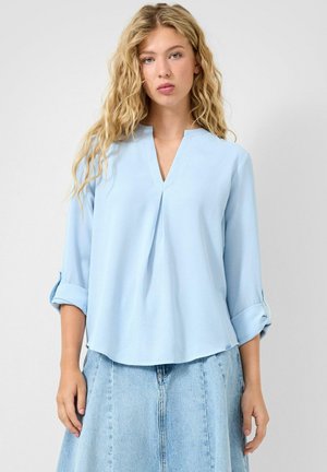 Femme blonde aux boucles lâches portant un chemisier bleu clair à col en V avec manches retroussées et une jupe en denim clair taille haute, sur un fond uni.