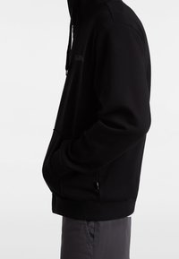 Person mit schwarzem Hoodie und Händen in der Fronttasche, kombiniert mit dunkelgrauer Hose, seitlich vor einfachem Hintergrund gezeigt.