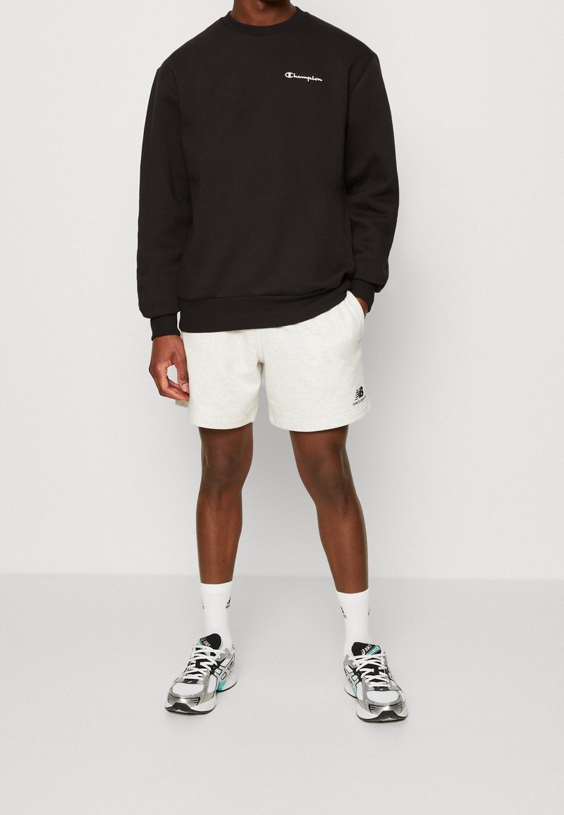 Homme portant un sweat-shirt noir Champion, un short blanc New Balance, des chaussettes blanches et des chaussures de course argentées, debout devant un fond uni.
