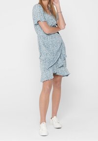 Blue floral dress with short sleeves, gathered detail at the side, ruffled hem, made from lightweight fabric, paired with white sneakers.