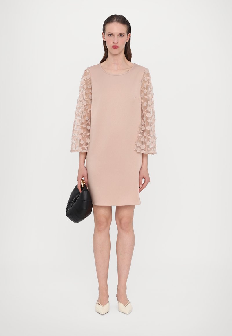 Robes de couleur rose avec des manches transparentes brodées de fleurs. Tissu mat, longueur genou, associée à un sac à main noir et des chaussures beiges à talons pointus.