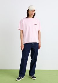 Pink short-sleeve t-shirt with "Dickies" logo, paired with dark blue denim pants and black sneakers. Model wears a beige cap.
