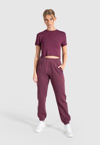 Bordeaux actiefkleding set met een cropped t-shirt met korte mouwen en losse joggerbroeken met zijzakken. Glad, rekbaar materiaal.