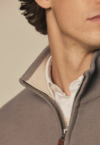 Grå zip-up sweater med ribbet tekstur, der har en cremefarvet indvendig krave og en hvid undertrøje med knapdetaljer.