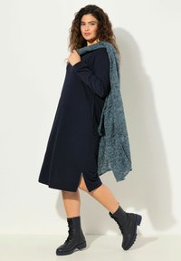 Laivastonsininen pitkähiha-asu, jossa on sivuvenytys, yhdistettynä teal-väriseen huiviin. Malli käyttää mustia nyörikenkäbootseja. Pehmeä kangasrakennus, rento istuvuus.