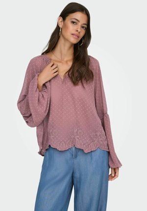 Vrouw in een mauve blouse met lange mouwen, een gestructureerd stippenpatroon en bloemenborduursel, gecombineerd met losse lichtblauwe broek.