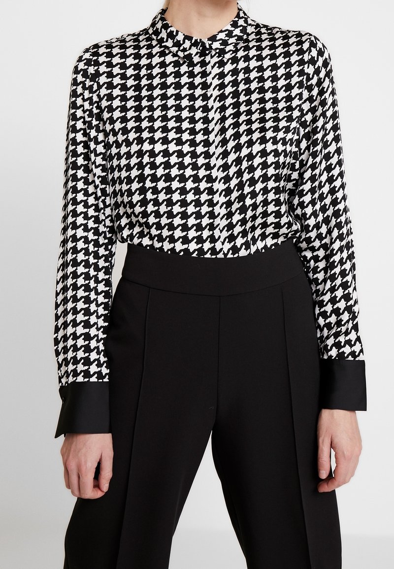 Femme portant une chemise à manches longues à motif pied-de-poule noir et blanc rentrée dans un pantalon noir taille haute avec de larges poignets.