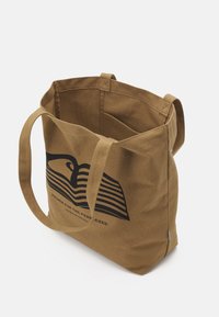 Ruskea puuvillainen kangaskassi, jossa musta graafinen kuvitus, mukaan lukien aaltoilevat viivat ja teksti, varustettu kahdella pitkällä kahvalla ja avoimella yläosalla.