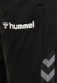 Svarta träningsbyxor med en vit "hummel"-logotyp, strukturerad tyg och ett distinkt svart-vitt chevronmönster på sidan.