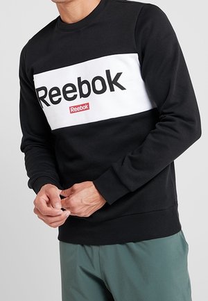 Mann trägt schwarzen und weißen Reebok-Sweatshirt mit Logo auf der Brust und grüne Hose und zieht seine Ärmel hoch.
